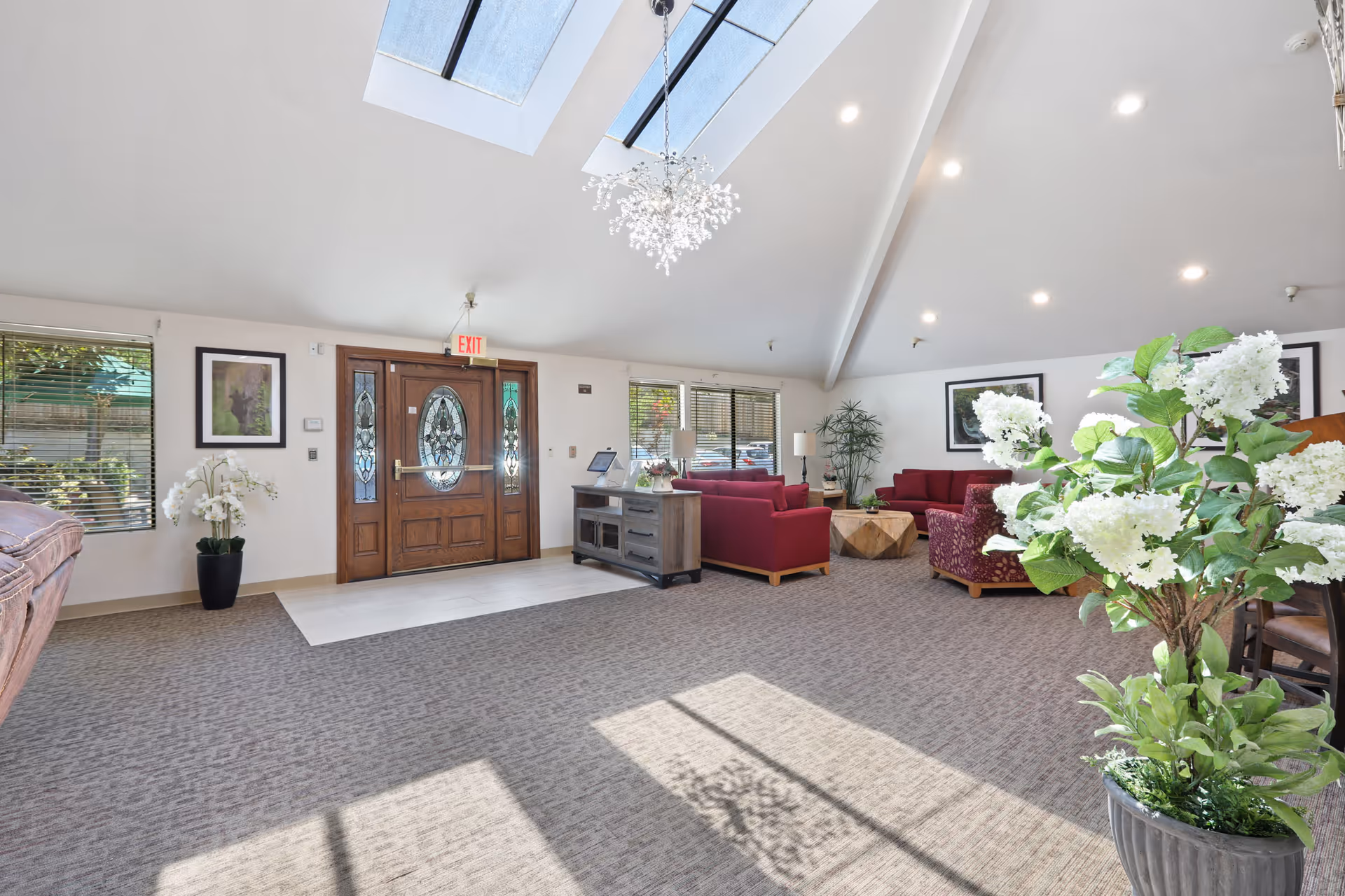 Bright and spacious assisted living facility common area with a high vaulted ceiling and skylights. The room features a wooden double door entrance with decorative glass panels, several red upholstered chairs and sofas arranged around a wooden coffee table, potted plants including a large floral arrangement in the foreground, framed artwork on the walls, and large windows letting in natural light.