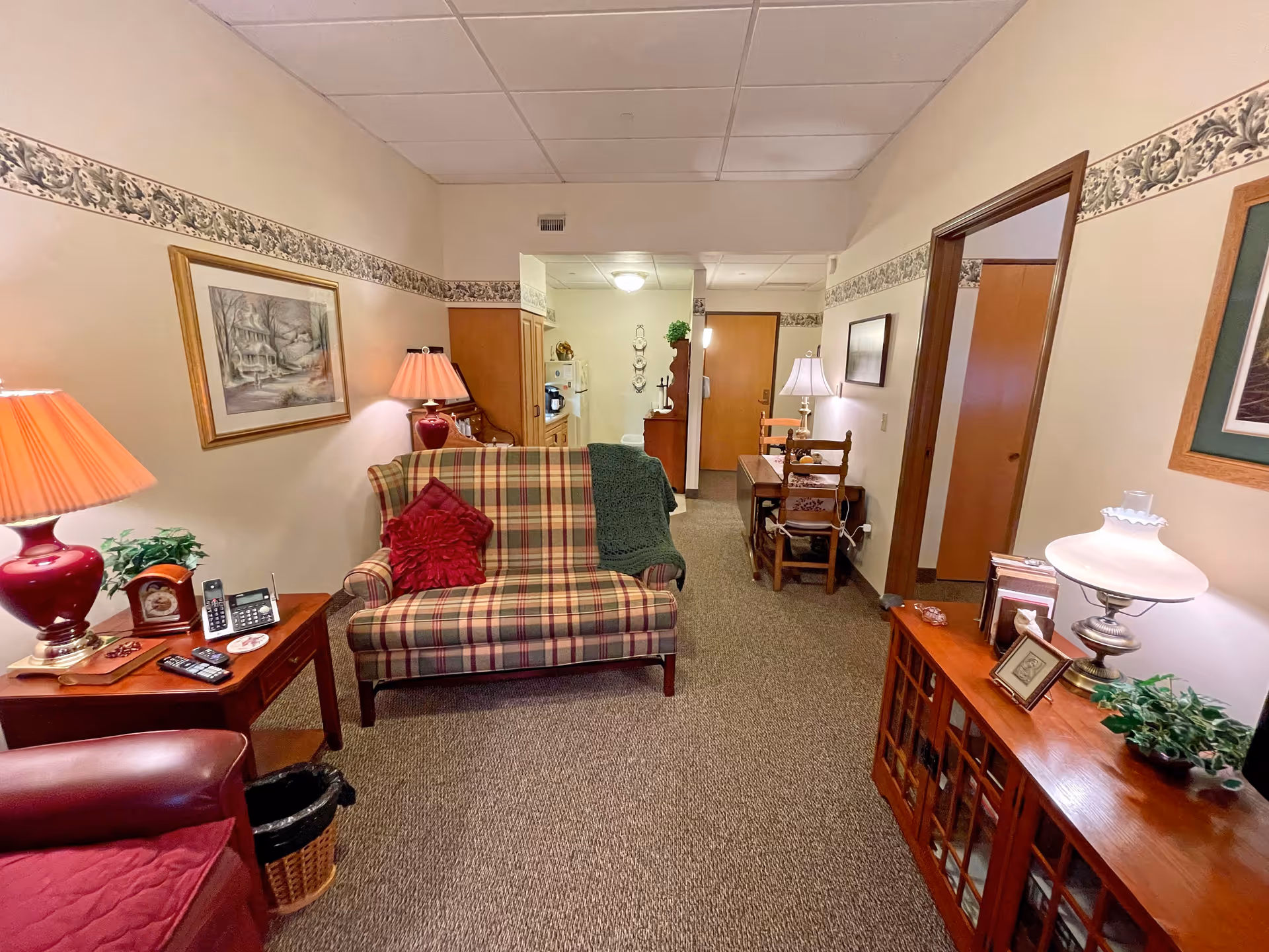 A cozy living room with a plaid loveseat, lamps, side tables and wooden cabinets opening to a hallway.