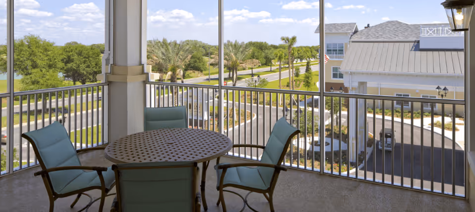 Screened balcony with a round table and four teal chairs overlooking the facility grounds, driveway, and neighboring building.