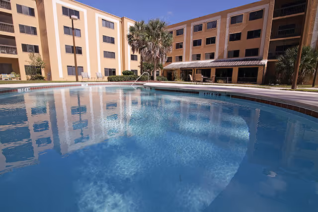 Outdoor swimming pool in front of a multi-story senior living facility building with palm trees and clear blue sky.