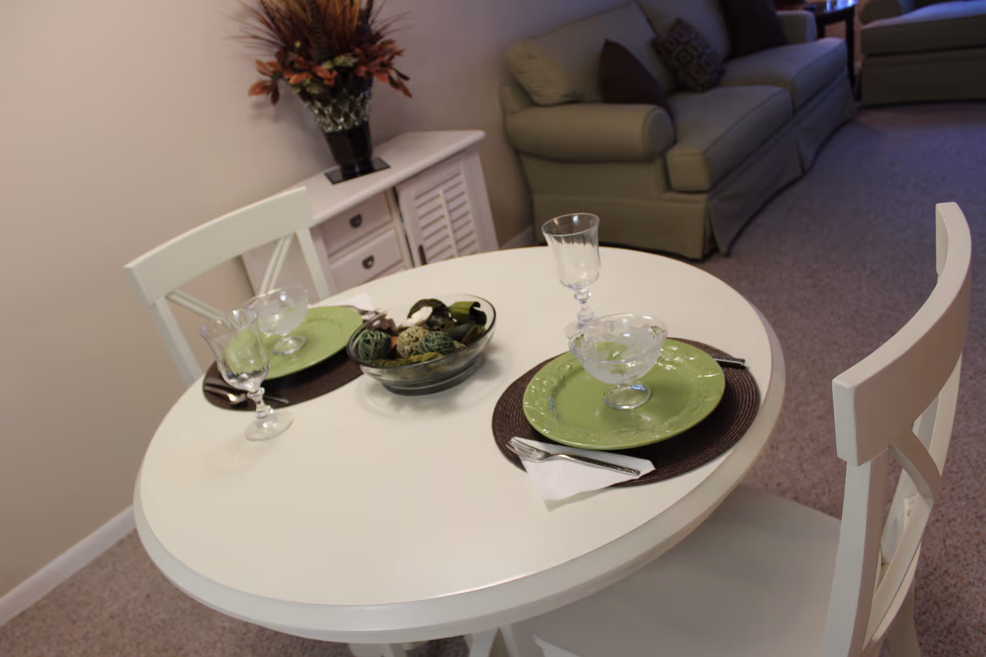 A small round white dining table set for two with green plates, glassware, and silverware on dark placemats. A decorative bowl with assorted natural elements is in the center. In the background, there is a white sideboard with a vase of dried flowers and a beige sofa in a carpeted room.