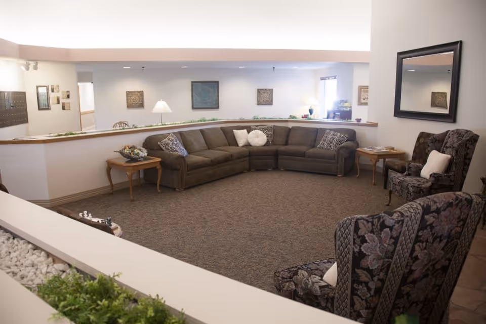A cozy living room area in a senior living facility featuring a large brown sectional sofa with decorative pillows, two floral patterned armchairs with white cushions, wooden side tables, a floor lamp, framed artwork on the walls, and a large mirror. The room has carpeted flooring and a half wall with plants on top.