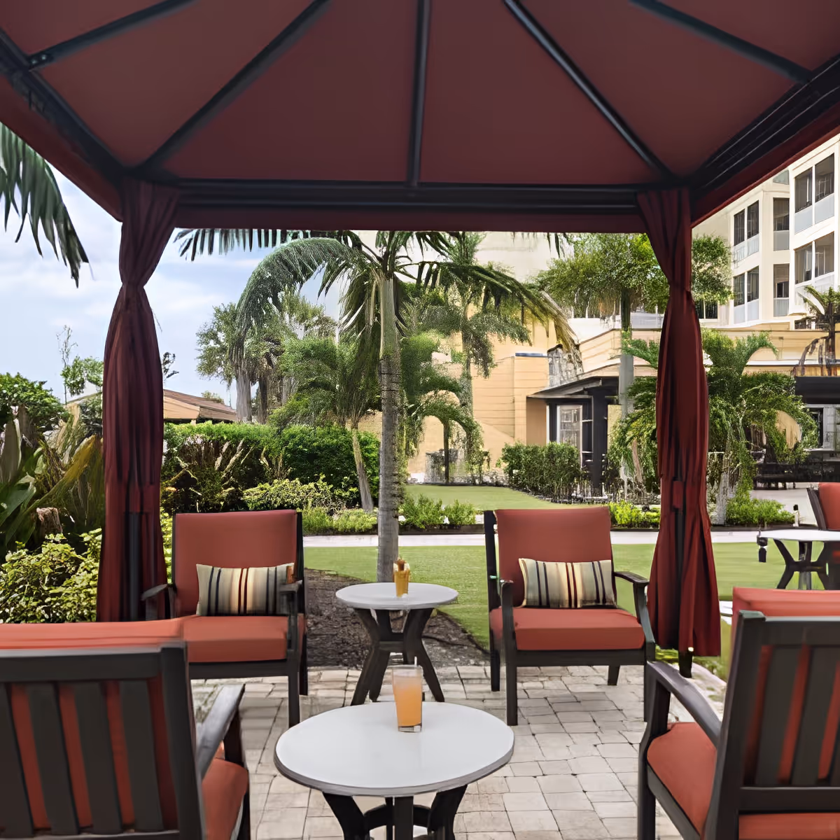 Outdoor seating area under a red canopy with cushioned chairs and small round tables, surrounded by greenery and palm trees, with a building visible in the background.