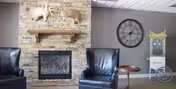 Cozy living room area with a stone fireplace featuring mounted animal heads above the mantel, two black leather armchairs, a small wooden side table, and a large wall clock on a gray wall.