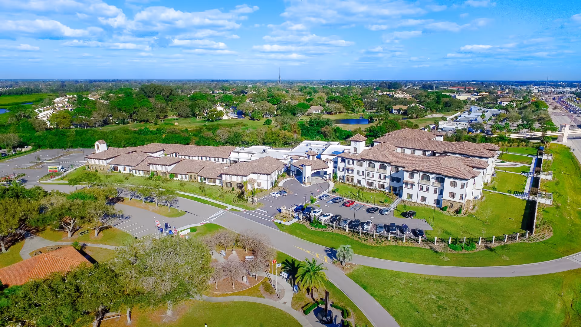 Aerial view of Tuscan Gardens Of Venetia Bay senior living complex with Mediterranean-style buildings, circular driveway, parking lot and surrounding green lawns.