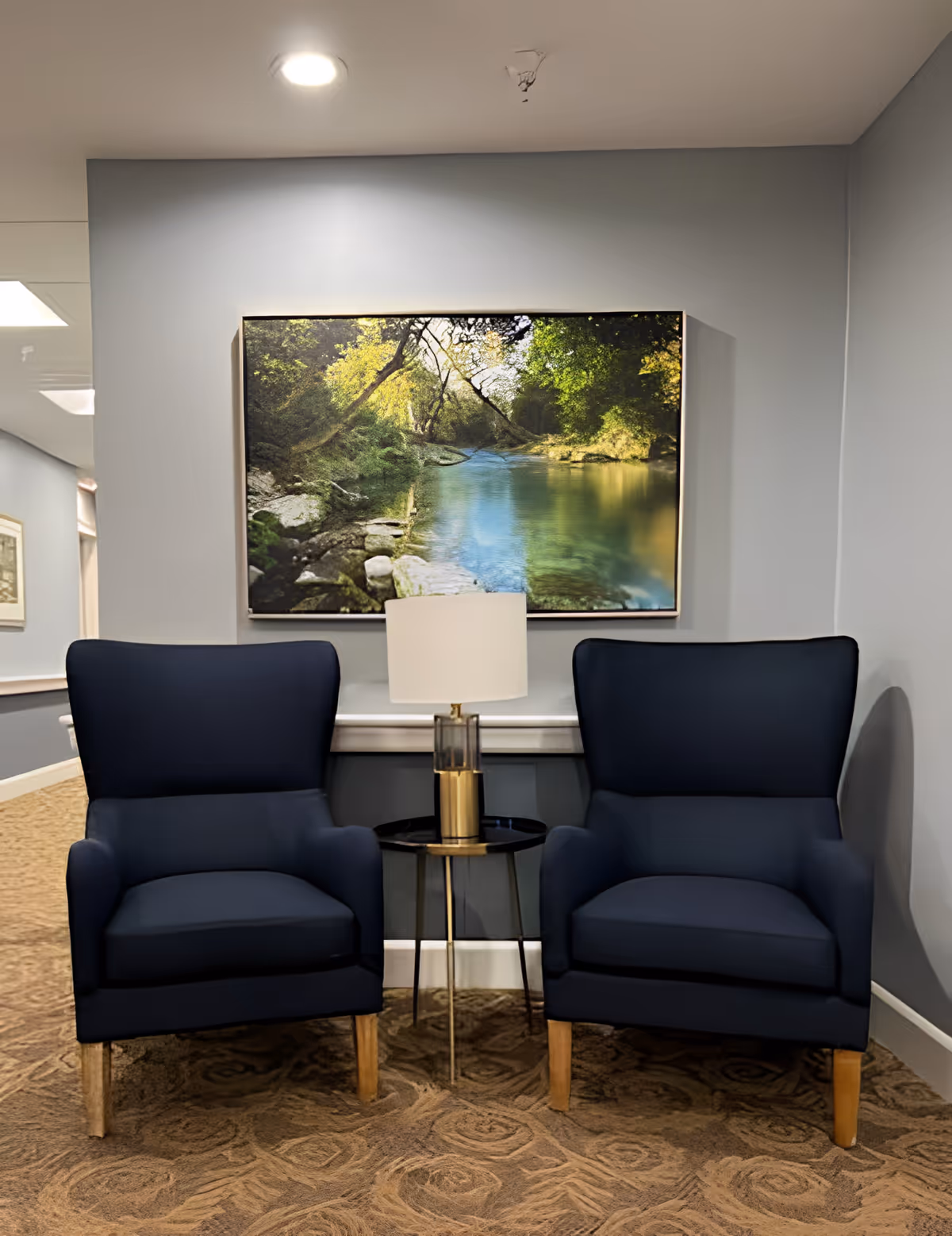 Two dark blue armchairs with wooden legs are positioned side by side in a hallway or common area with a small round black side table between them holding a lamp with a white shade. Above the chairs hangs a large framed picture of a serene river scene with trees and rocks. The walls are painted light gray and the floor is carpeted with a patterned design.