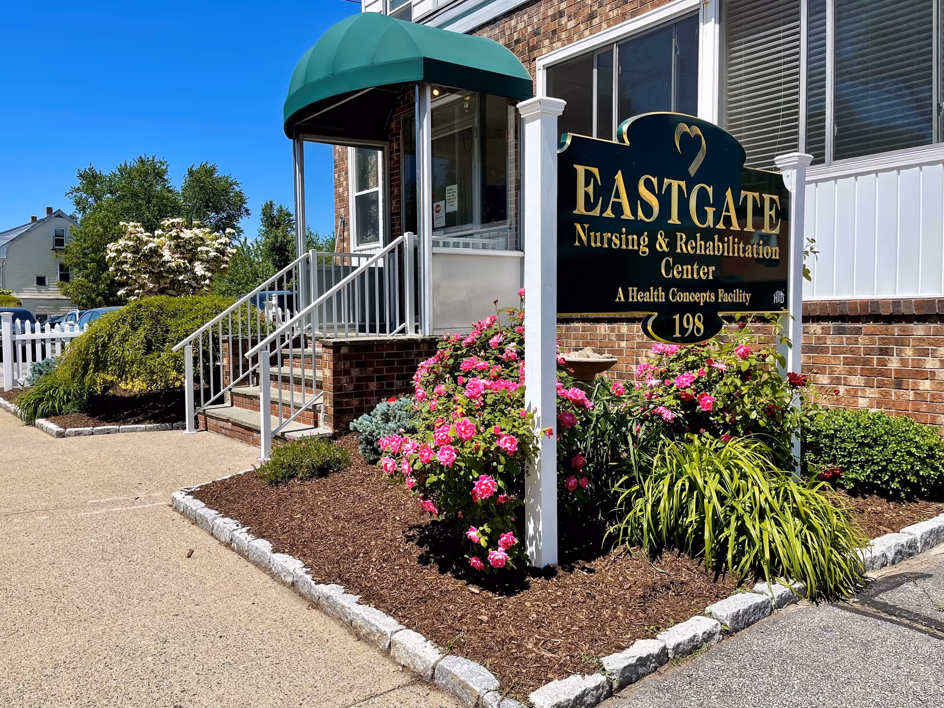 Entrance of Eastgate Nursing & Rehabilitation Center with a green awning over the doorway, a sign displaying the facility name, and a landscaped garden with pink flowers and greenery.