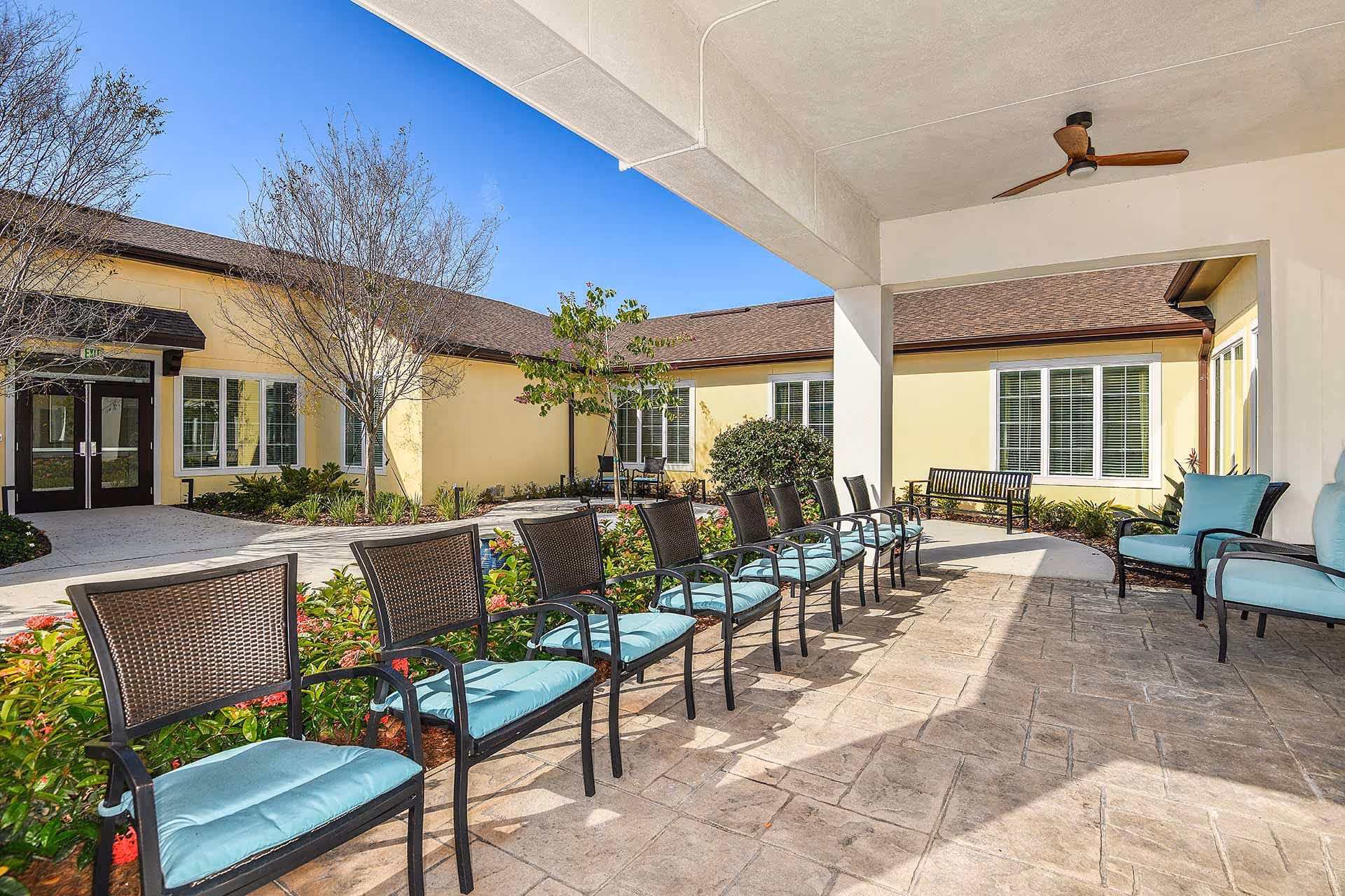 Outdoor covered patio area at Providence Living at Hunter's Creek with a row of black metal chairs with light blue cushions, a ceiling fan, and a view of the building with yellow walls and multiple windows under a clear blue sky.