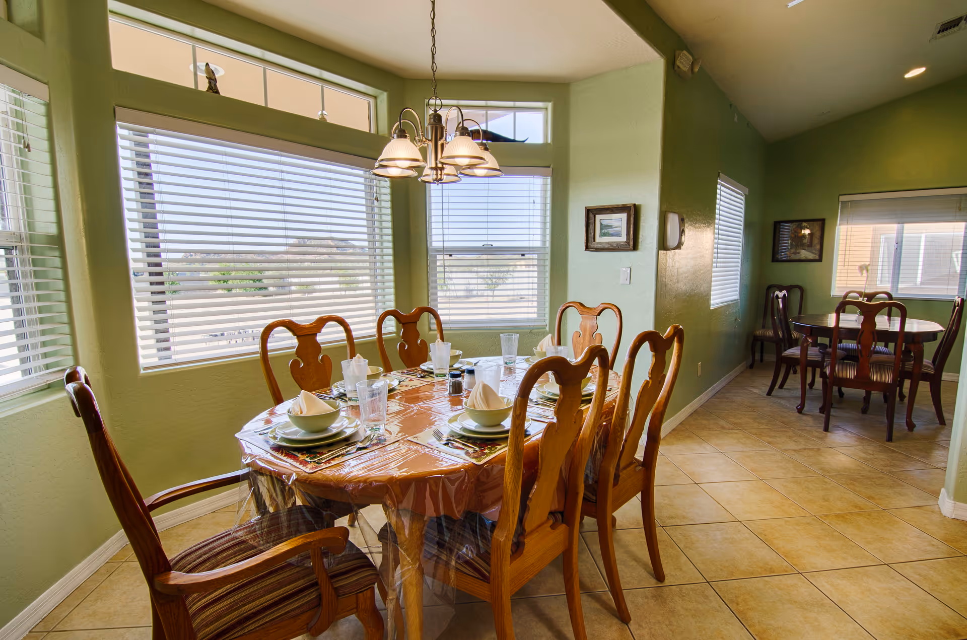 A bright dining room with green walls and large windows with white blinds. There is a wooden dining table covered with a clear plastic tablecloth, set with plates, bowls, napkins, and glasses. Six wooden chairs with cushions surround the table. In the background, there is another smaller dining table with four chairs. The floor is tiled, and a chandelier hangs above the main dining table.