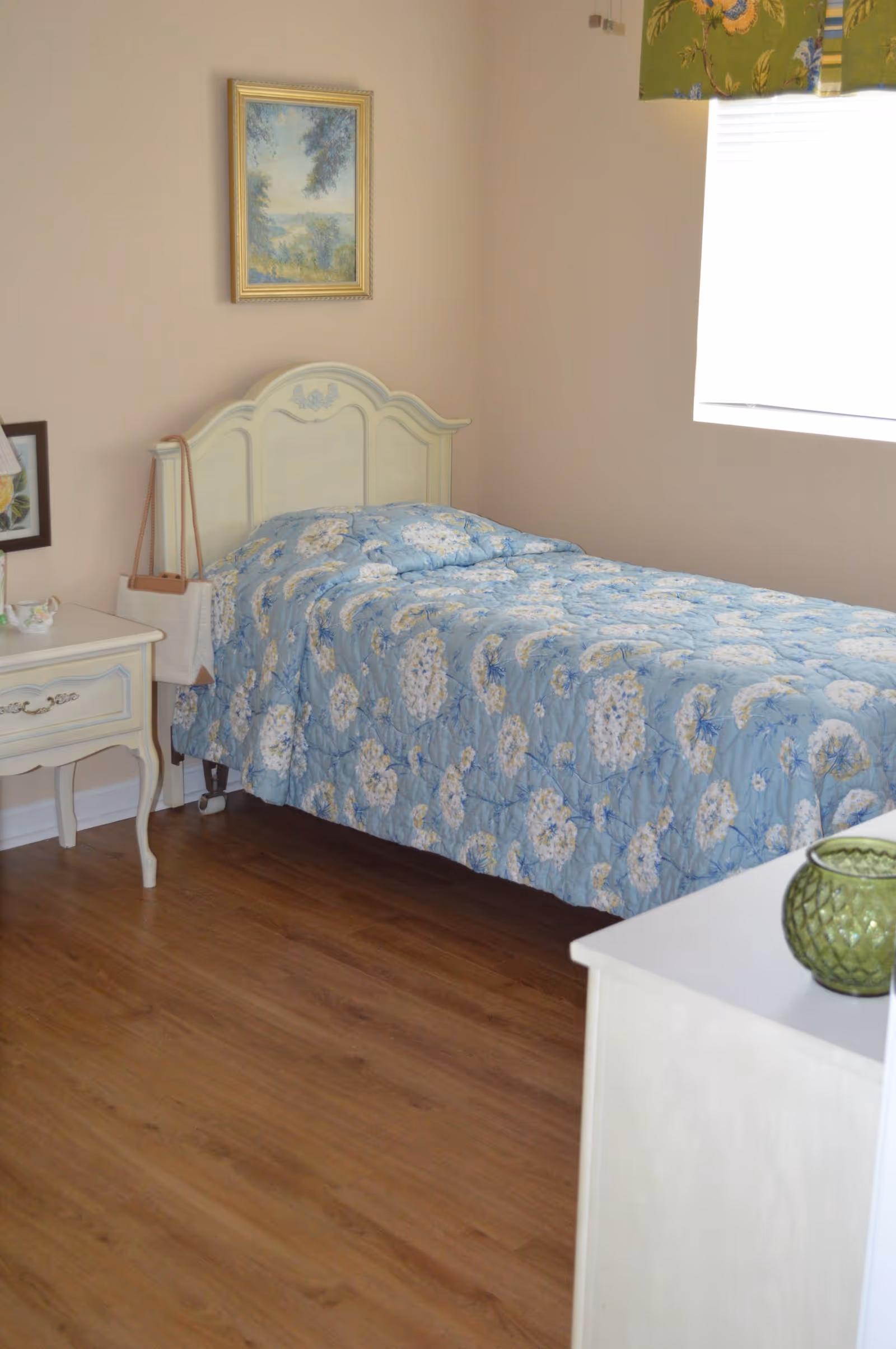 Sunlit bedroom with a single bed covered in a blue floral quilt, a nightstand, framed art, and a window.