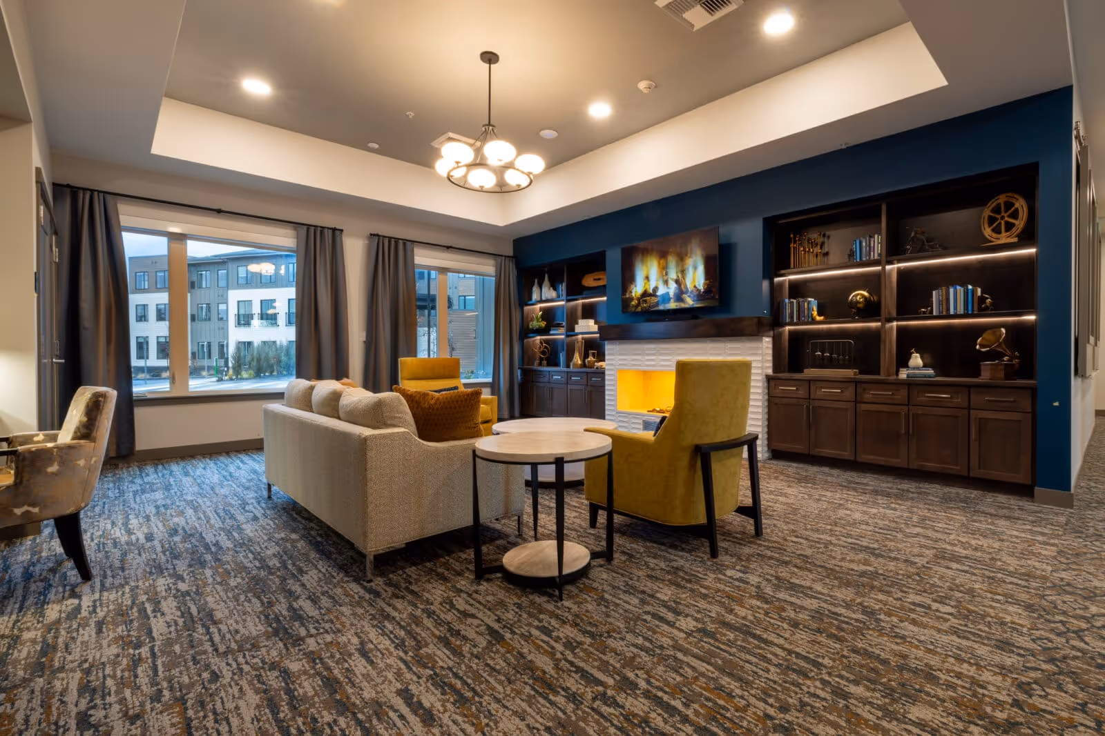 A spacious senior living lounge with sofas, yellow armchairs, a fireplace beneath a mounted TV, built-in shelving, and large windows.