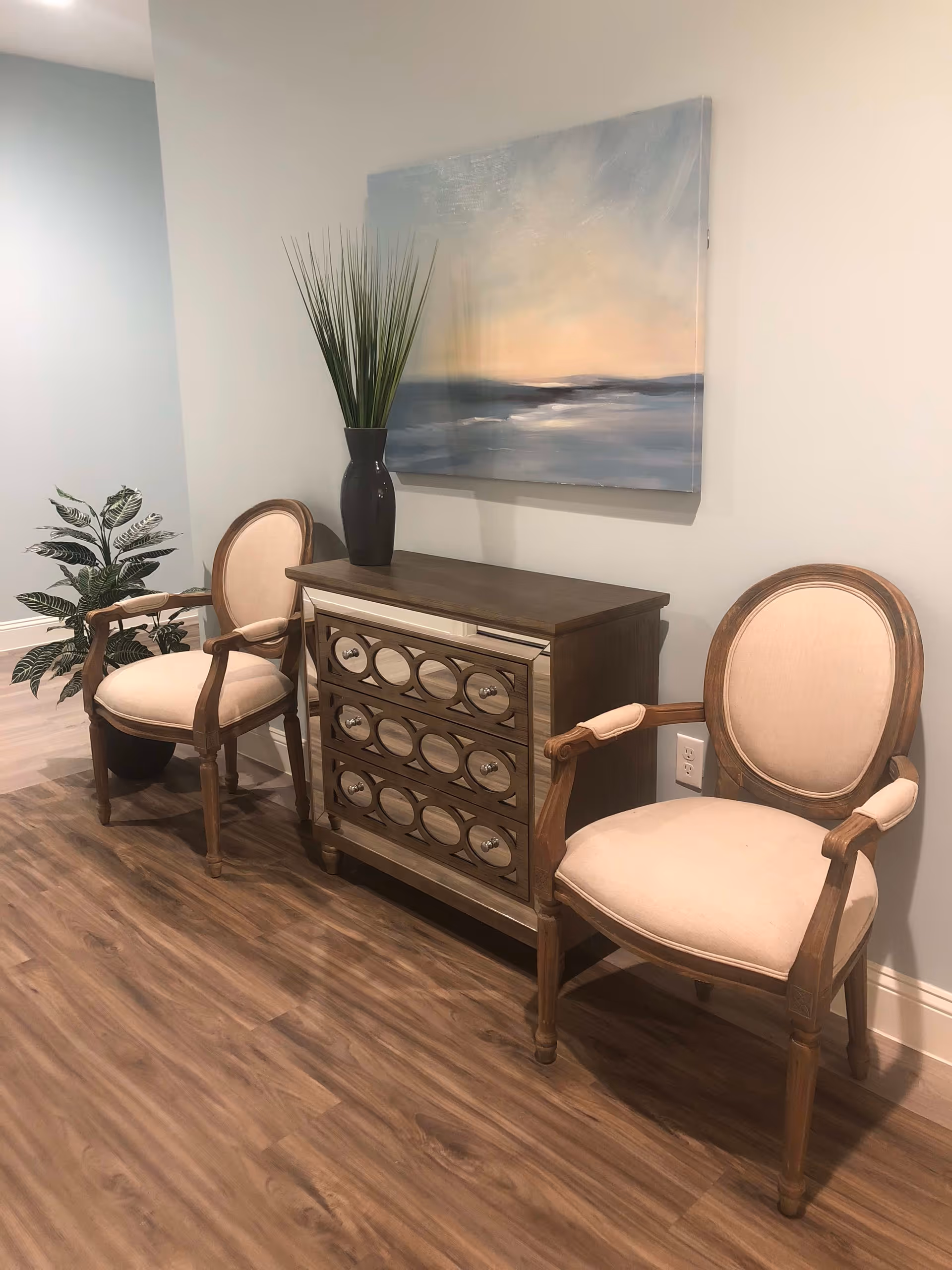 Two wooden armchairs with beige cushions are placed on either side of a wooden chest of drawers with circular mirror accents. A tall black vase with green decorative grass sits on top of the chest. A large painting depicting a serene landscape with soft blue and orange hues hangs on the wall above the chest. A potted plant is visible in the corner of the room with light-colored walls and wood flooring.