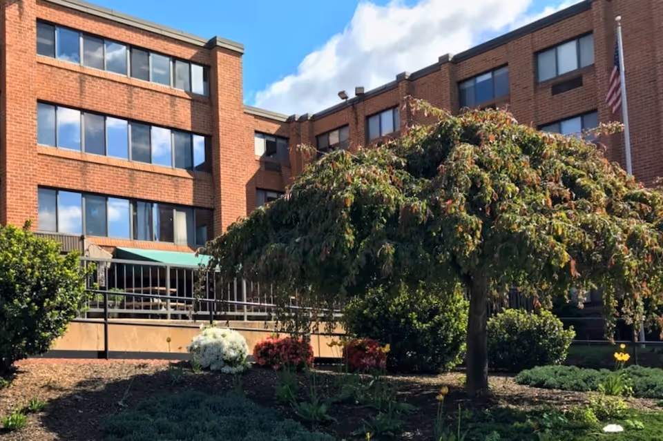 Brick multi-story building with rows of windows, a landscaped garden and a leafy tree in front under a blue sky.