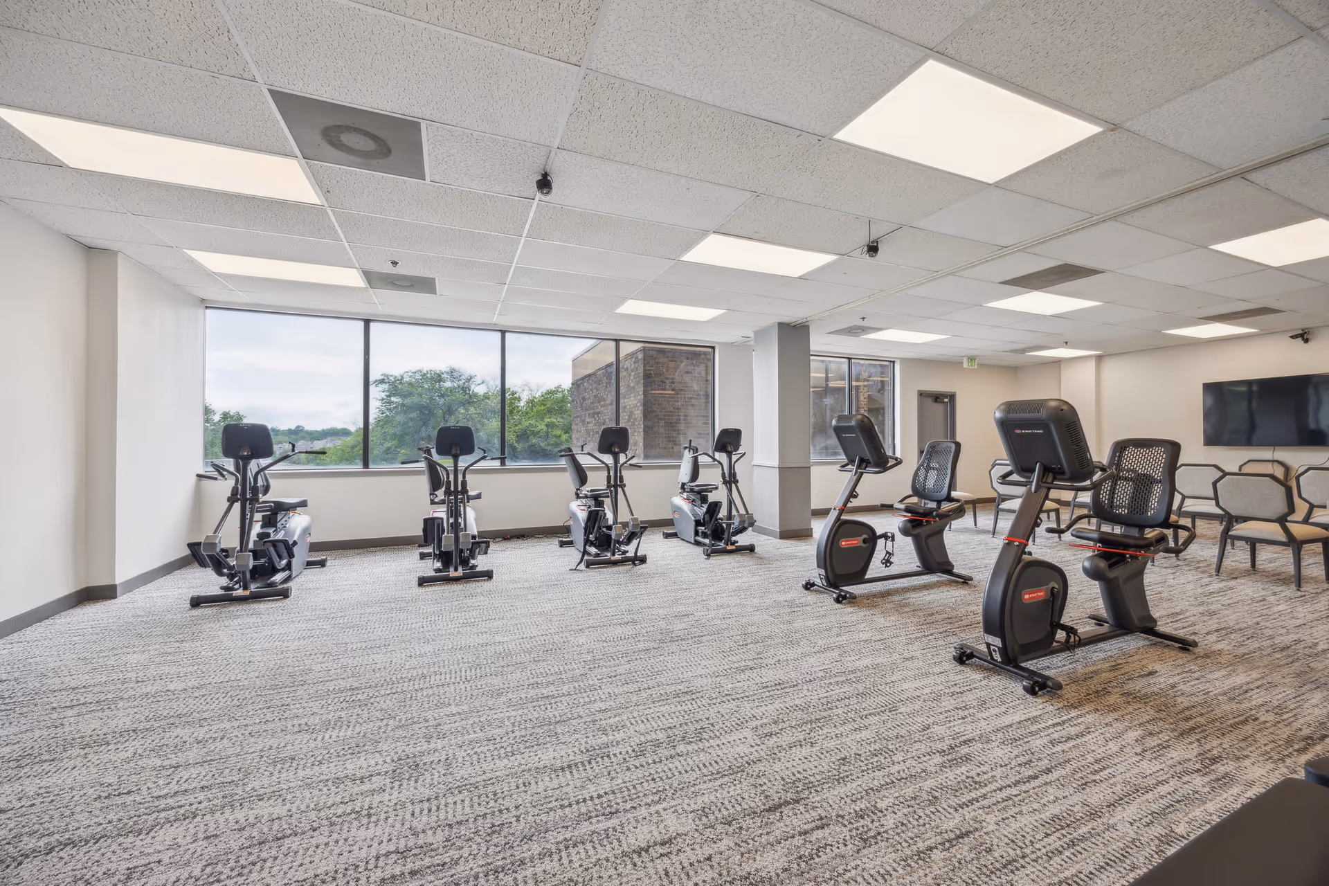 A spacious exercise room with several stationary recumbent bikes and elliptical machines arranged on a patterned carpet. Large windows on one side provide natural light and a view of trees and a building outside. The ceiling has recessed lighting panels, and there is a wall-mounted TV with chairs arranged nearby.