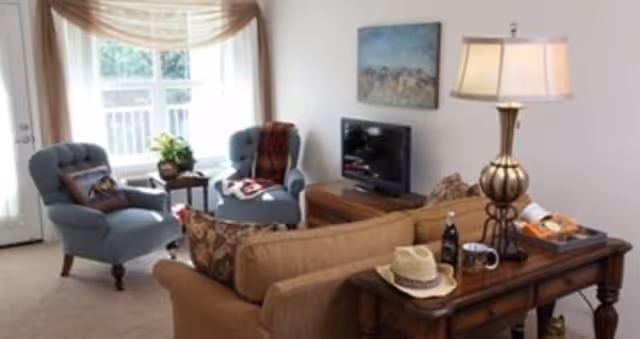 A cozy living room with a beige sofa, two blue armchairs, a wooden side table with a lamp, a small TV on a wooden stand, and a window with sheer curtains letting in natural light.