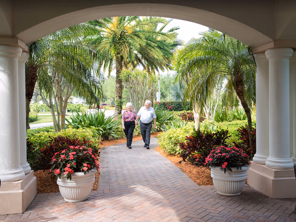 An older couple walks together along a landscaped brick walkway framed by columns, potted flowers, and palm trees.