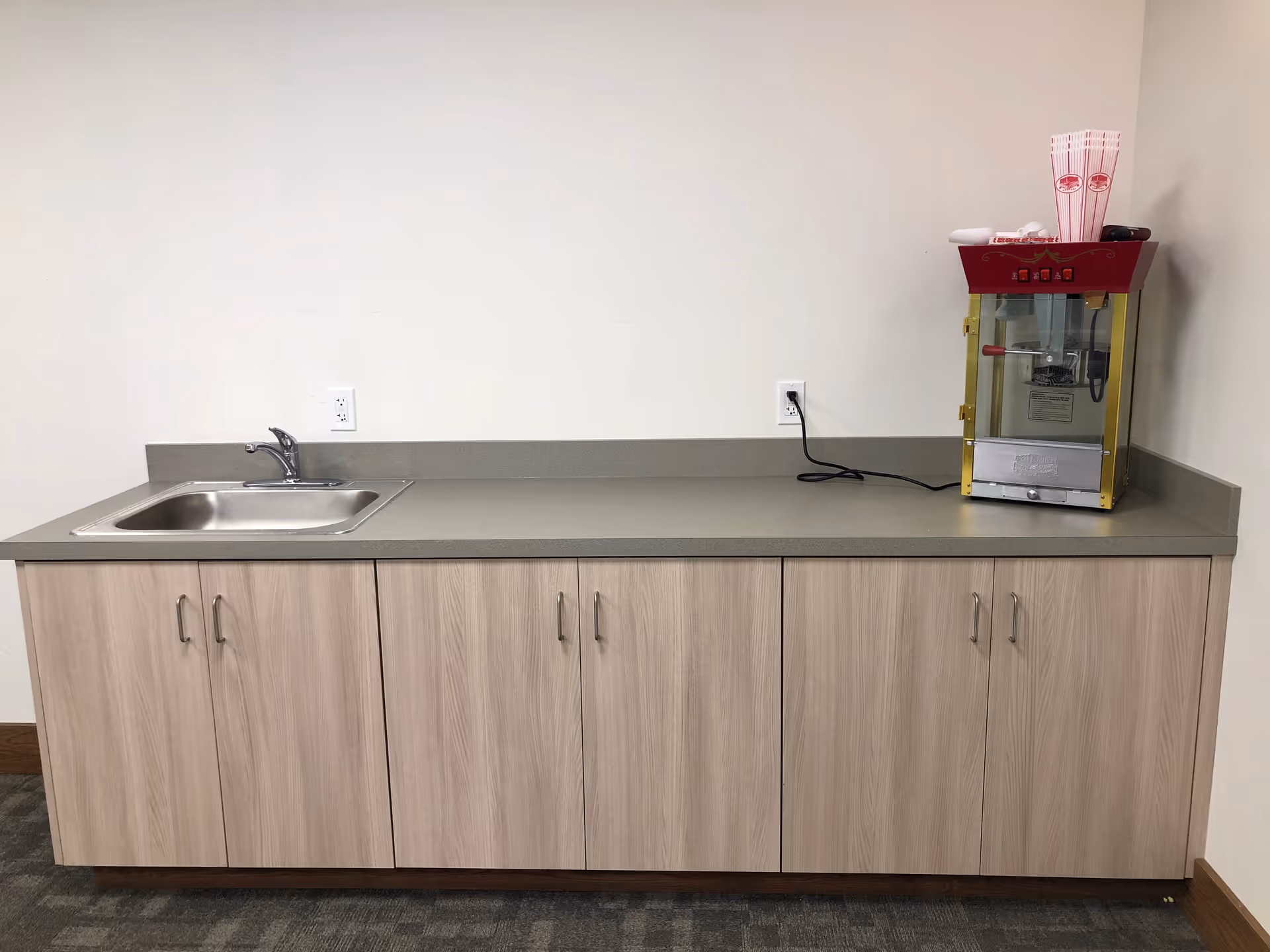 A kitchenette countertop with a sink, wood cabinets below, and a popcorn machine on the right against a plain wall.