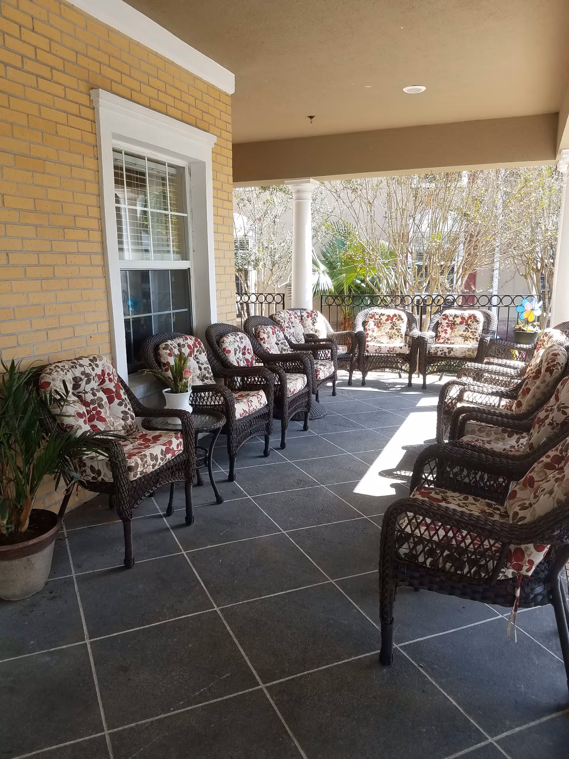 Covered outdoor patio area with multiple wicker chairs arranged in two rows facing each other. The chairs have floral patterned cushions. There are potted plants on the tiled floor and a yellow brick wall with a window on the left side. White columns support the roof of the patio, and trees and shrubs are visible outside the railing.