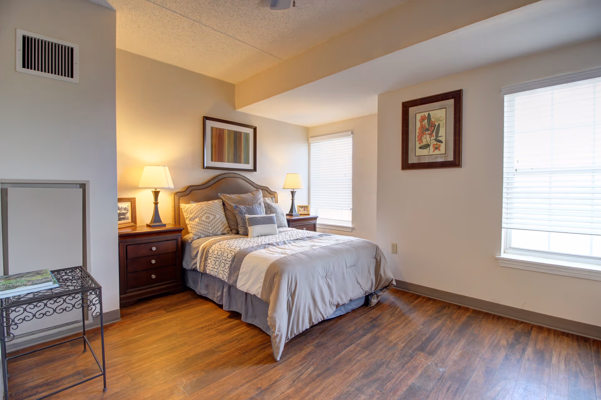 A neatly made bed with multiple pillows in a senior living bedroom. The room has wooden floors, two bedside tables with lamps, framed artwork on the walls, and two windows with blinds letting in natural light.