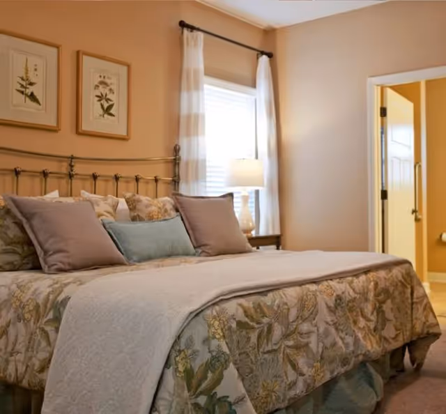 Cozy bedroom with a floral bedspread, multiple pillows, a bedside lamp, and framed botanical prints on the wall.