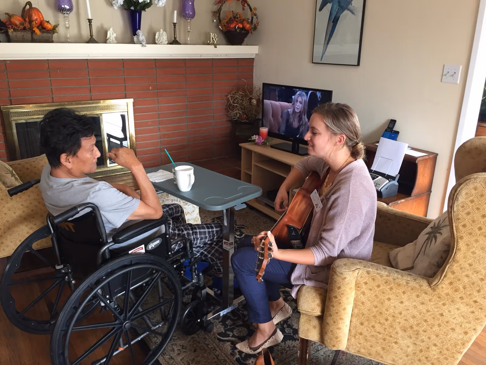 A woman plays guitar for a man in a wheelchair in a cozy living room with a fireplace and TV.
