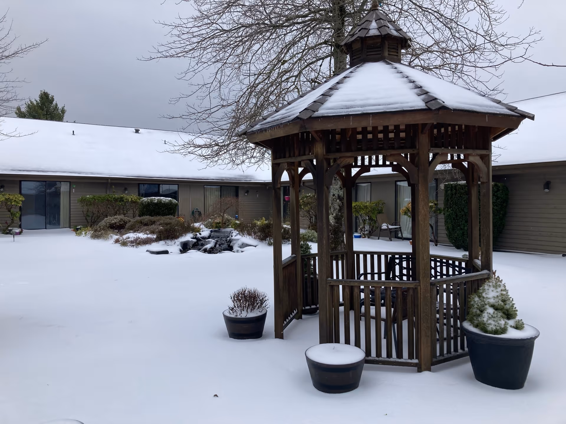 Snow-covered courtyard at Channel Point Village featuring a wooden gazebo with a snow-covered roof, surrounded by potted plants and bushes. The building with multiple windows and sliding doors encloses the courtyard.
