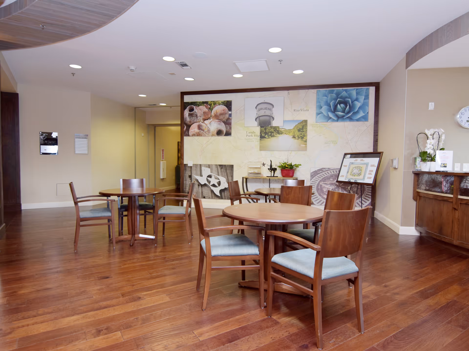 A common area with wooden floors and several round wooden tables surrounded by chairs with blue cushions. The walls are decorated with a large mural featuring various images including pottery, a water tower, a succulent plant, and a map. There is a small table with a plant and framed artwork on the right side, along with a clock on the wall and a wooden cabinet below it.