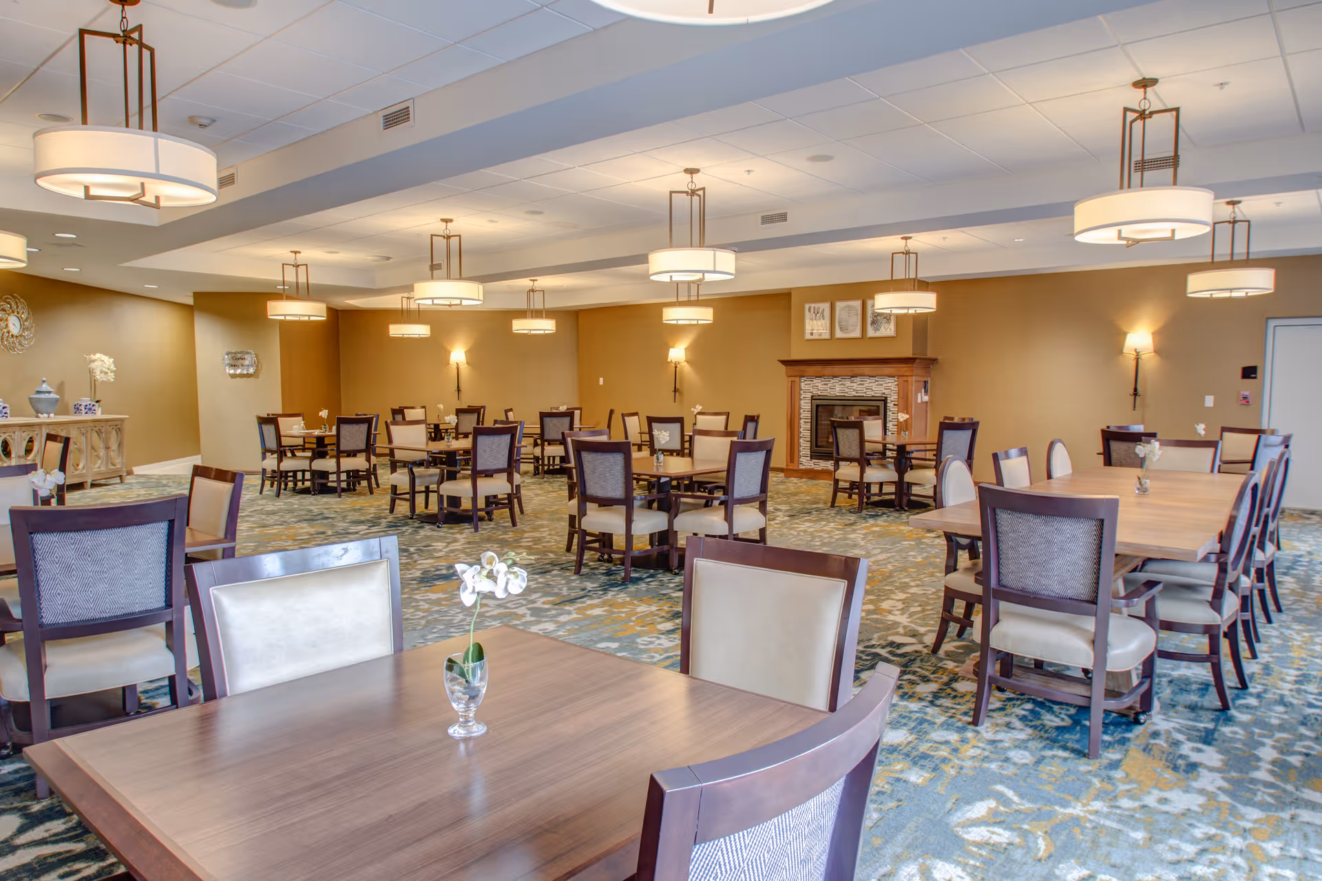 A spacious dining room with multiple wooden tables and chairs arranged neatly. The room features a patterned carpet, beige walls, modern hanging light fixtures, and a fireplace with framed artwork above it. Small flower vases are placed on the tables.