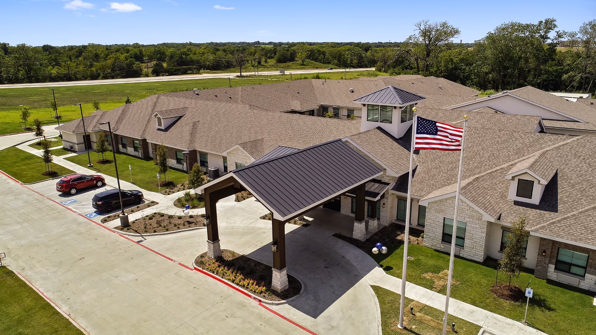 Aerial view of Celebration Senior Living of Denison building with a covered entrance, two flagpoles with an American flag, surrounding greenery, and a parking area with a few cars.