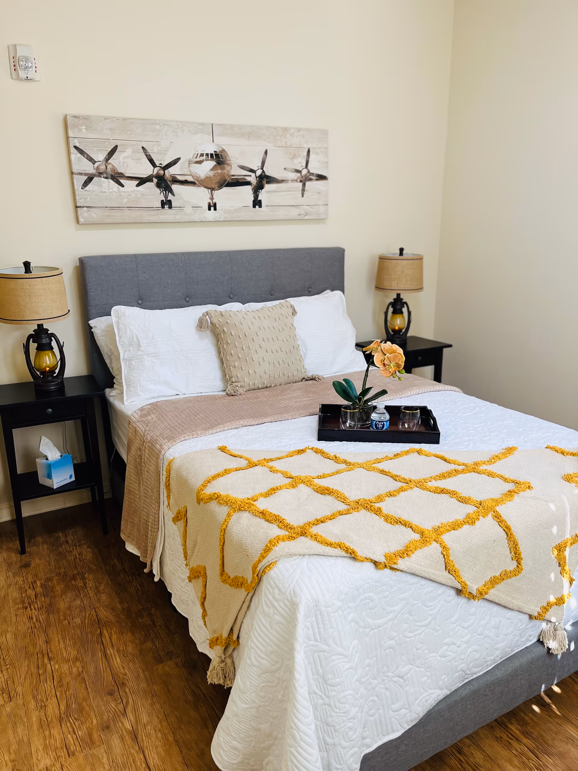 Sunlit bedroom with a neatly made bed featuring a gray headboard, yellow-accent throw, nightstands with lamps, and airplane wall art.