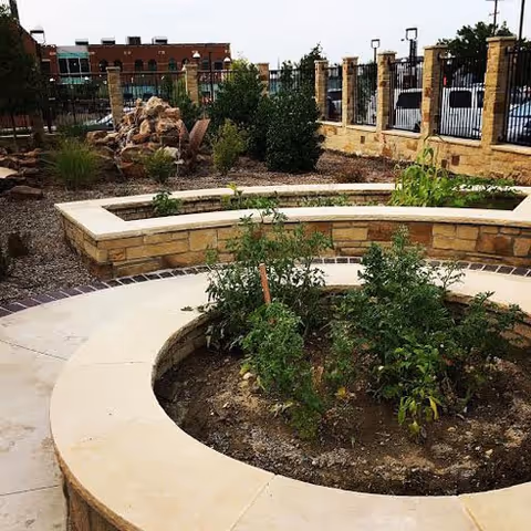 Outdoor garden area with circular raised stone planters containing green plants and shrubs, surrounded by a paved walkway and a stone fence with metal railings. In the background, there are buildings and trees.