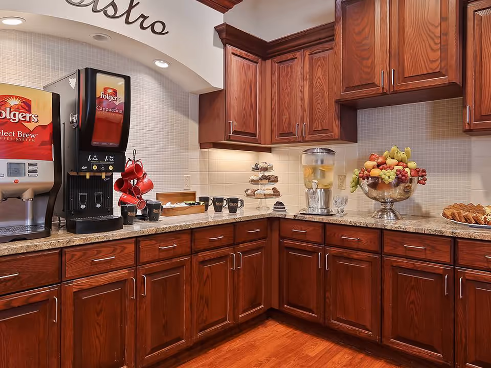A bistro-style kitchenette with wooden cabinets, coffee machines, a fruit bowl, and pastries on a granite countertop.