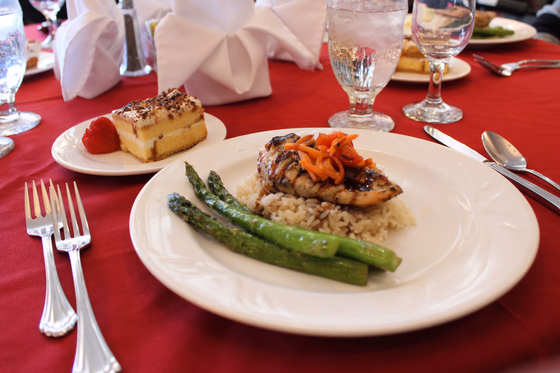 A plated meal on a white plate with grilled chicken topped with sautéed vegetables served over rice, accompanied by three asparagus spears. Next to the plate is a dessert plate with a slice of cake and a strawberry. The table is covered with a red tablecloth and set with silverware, water glasses, and folded white napkins.