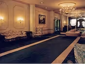 An elegant interior hallway or lobby area with classic decor including upholstered chairs, wall sconces, framed artwork, a chandelier, and a long carpet runner.