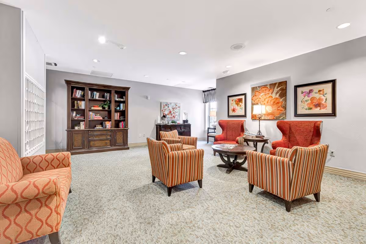 A bright and spacious living room area with patterned armchairs and a sofa arranged around a wooden coffee table. The walls are decorated with floral artwork, and a wooden bookshelf filled with books is visible in the background. The room has a light-colored carpet and recessed ceiling lights.