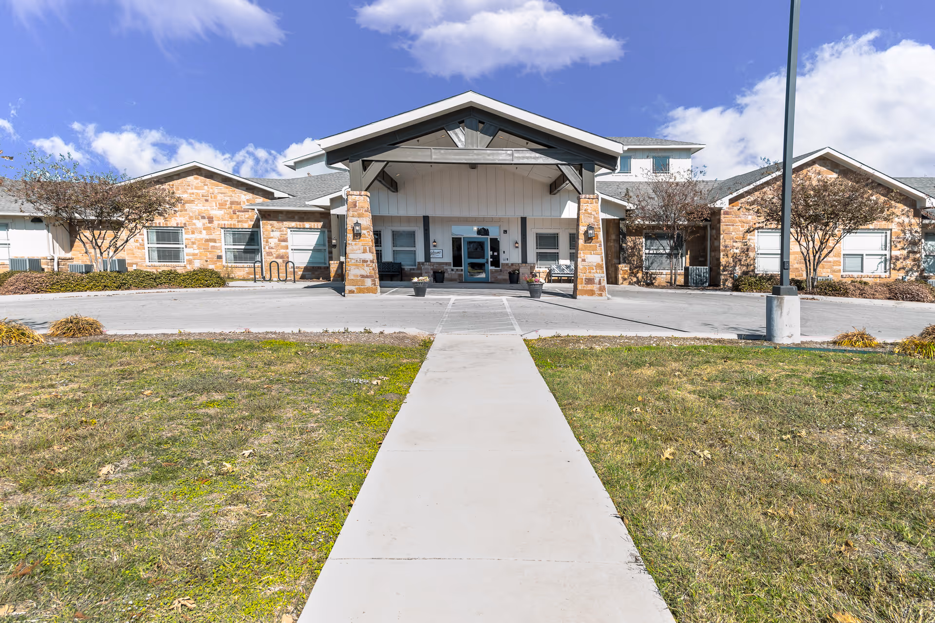 Front entrance of a single-story senior living facility with a concrete walkway leading to a covered entry flanked by stone facades and landscaping.