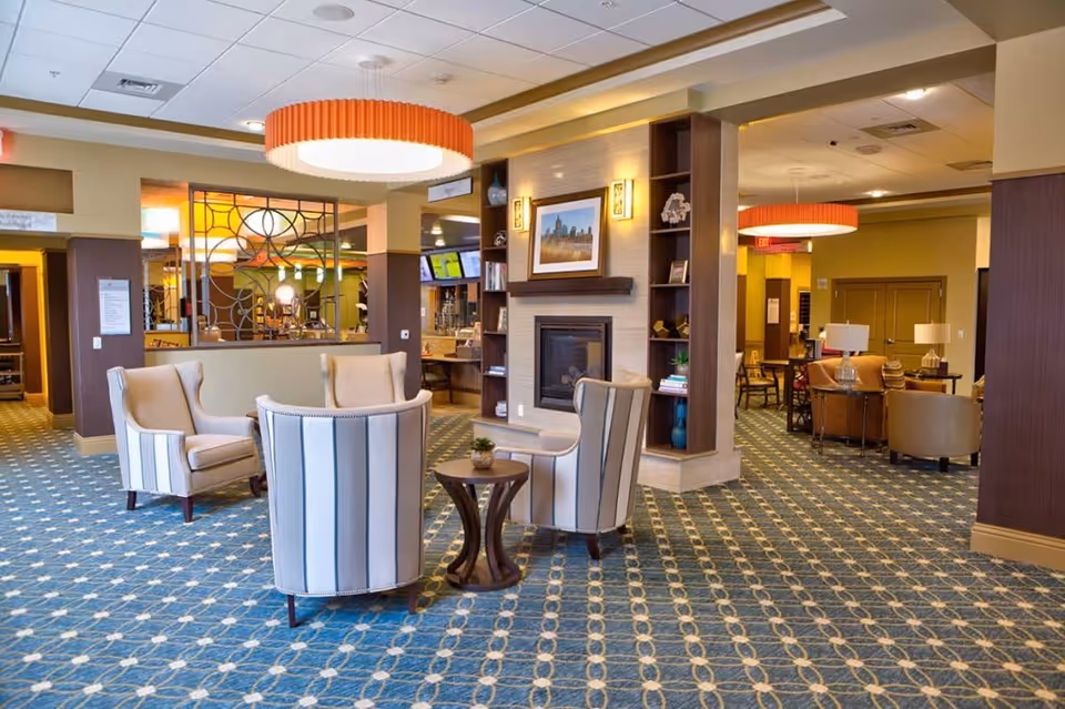 A spacious senior living community lounge area with patterned blue carpet, four striped armchairs arranged around a small wooden table, and a fireplace with built-in shelving. The room is warmly lit with large circular ceiling lights and decorated with framed artwork and various decorative items on shelves. Additional seating areas with chairs and tables are visible in the background.