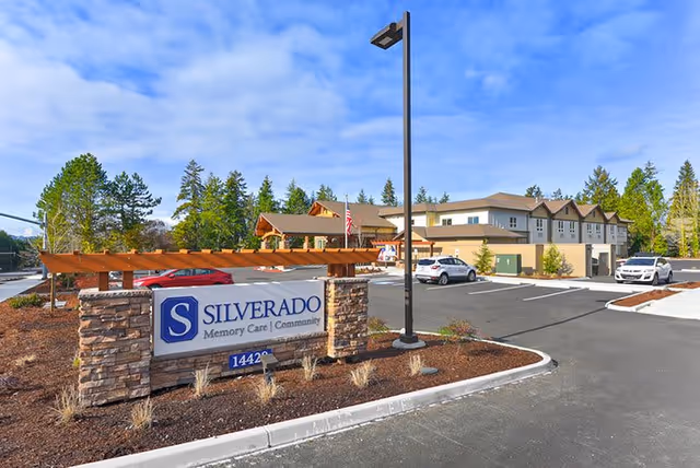 Exterior view of Silverado Bellevue Memory Care Community building with a parking lot in front, a stone sign displaying the facility name, and trees in the background under a partly cloudy sky.