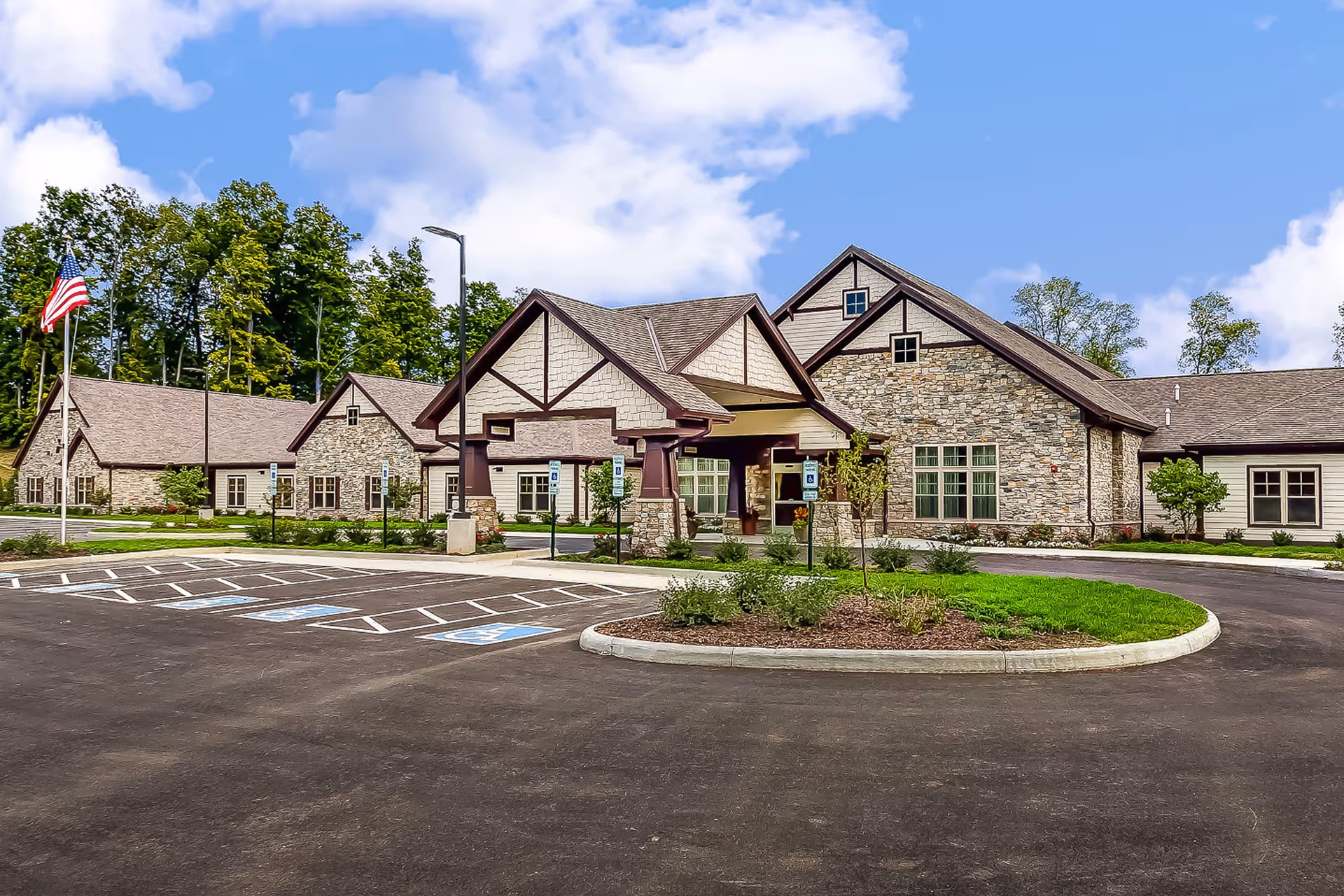 Exterior view of a senior living facility named Maplewood at Twinsburg, featuring a large stone and wood building with a covered entrance, surrounded by a parking lot with handicap spaces and landscaped greenery under a partly cloudy blue sky.