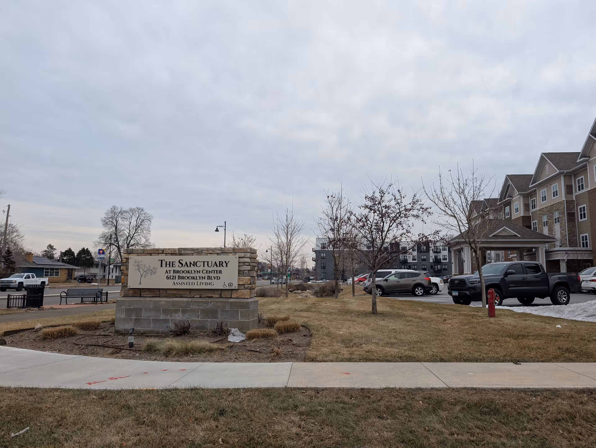 Outdoor view of The Sanctuary at Brooklyn Center assisted living facility sign made of stone and concrete, with the building and parking lot visible in the background under a cloudy sky.