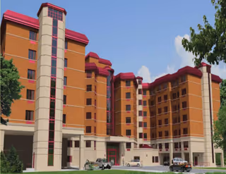 Exterior view of a multi-story assisted living facility building with red roofs and beige and orange walls, surrounded by trees and a few parked cars in front under a blue sky with some clouds.