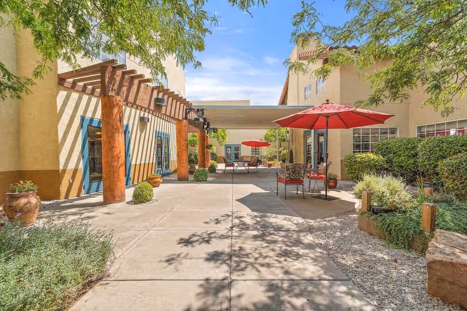 Sunny outdoor courtyard with patio tables and red umbrellas, a wooden pergola, and surrounding stucco buildings.