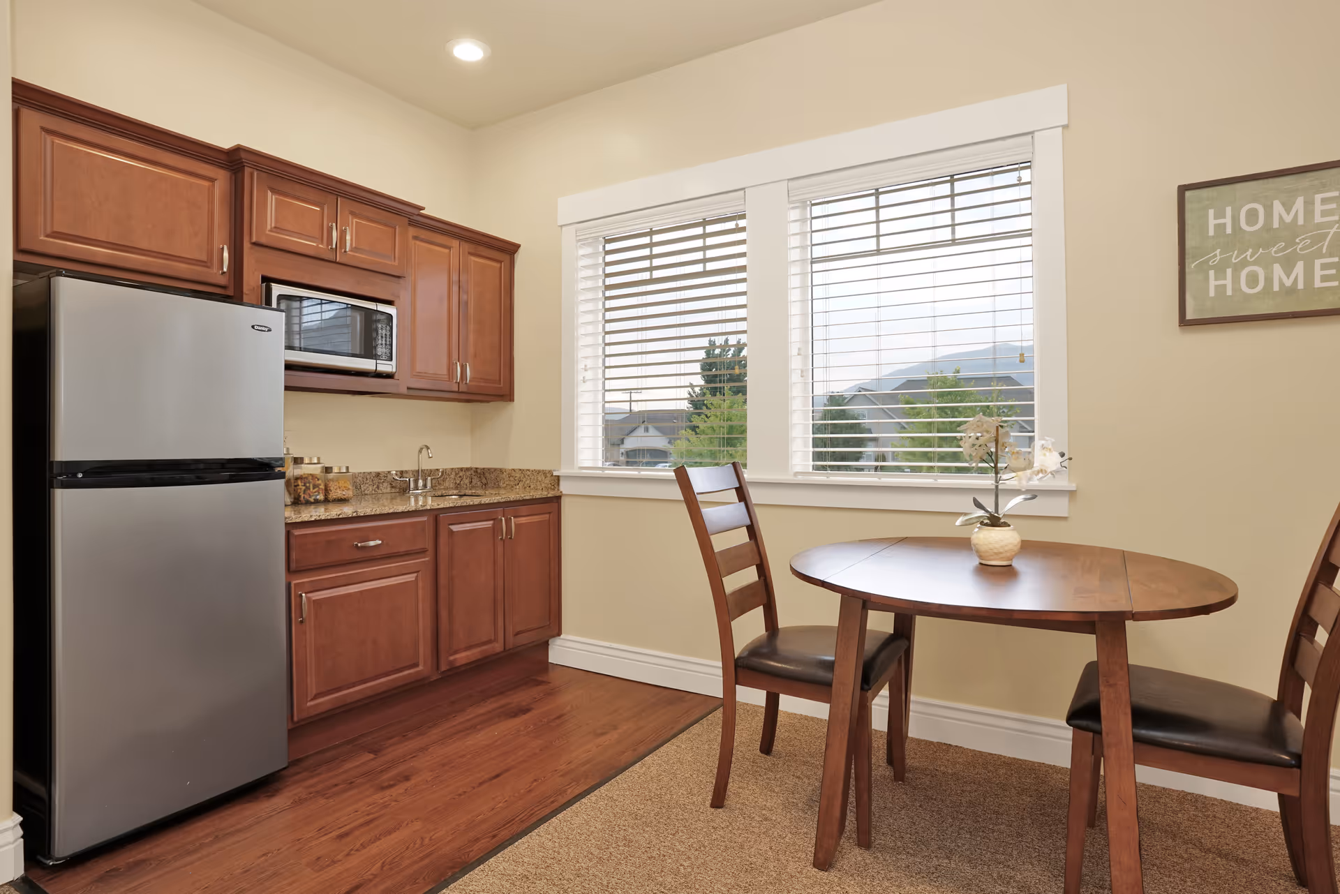 A small kitchen area with wooden cabinets, a stainless steel refrigerator, a microwave, and a granite countertop with a sink. Next to the kitchen is a wooden dining table with two chairs, a small vase with white flowers on the table, and a window with white blinds showing an outdoor view. A framed sign on the wall reads 'Home sweet Home'.