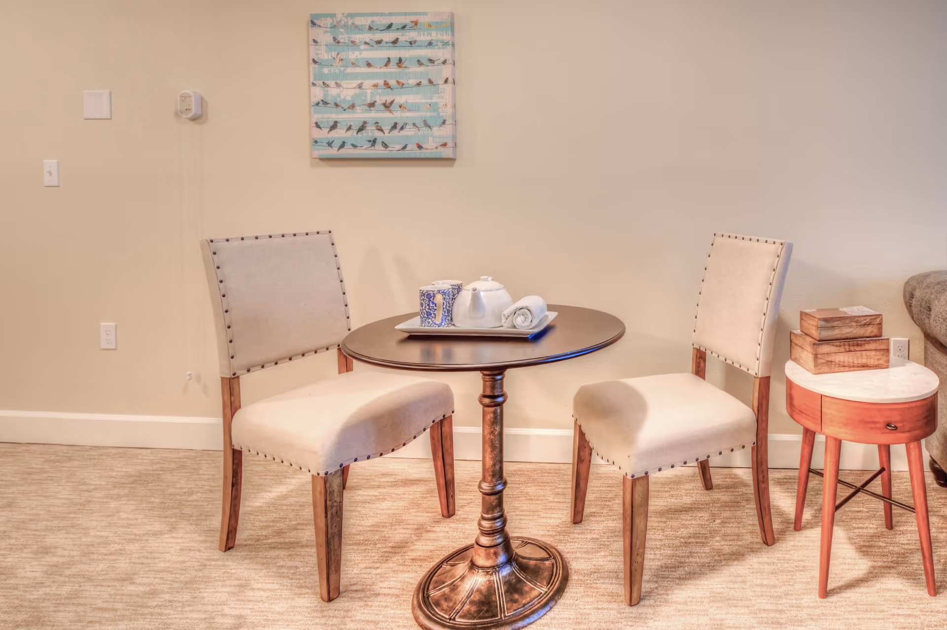 A small round wooden table with a teapot, cup, and rolled towel on a tray, flanked by two beige upholstered chairs with wooden legs. To the right is a small round side table with a marble top and two wooden boxes stacked on it. A wall-mounted artwork featuring birds on a blue background hangs above the table and chairs. The room has beige walls and carpeted flooring.