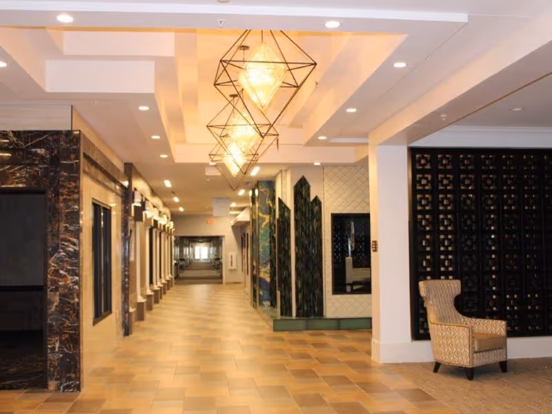 A spacious, well-lit hallway in a care and rehabilitation center with tiled floors, decorative wall panels, modern geometric pendant lights hanging from the ceiling, and a patterned armchair placed near a black lattice partition.