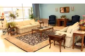 A bright living room with two beige sofas, a wooden coffee table on a patterned area rug, two dark blue armchairs, a wooden side table, and a large window letting in natural light. There are plants and decorative items including a snowman figure near the window.