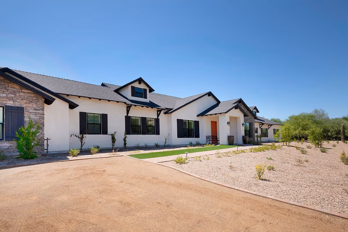 Exterior view of a single-story building with white walls, black window shutters, and a dark roof under a clear blue sky. The building is surrounded by a gravel landscape with sparse vegetation and a dirt driveway in front.