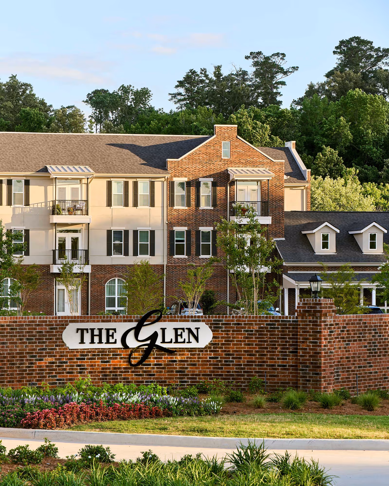 Exterior view of The Glen Retirement System building with a brick facade and multiple windows, surrounded by greenery and trees. A brick wall in front displays the sign 'THE Glen' with landscaped flowers and plants along the base.