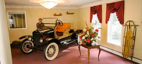 A vintage black car displayed indoors on a carpeted floor with a mannequin dressed as a driver seated inside. Next to the car is a wooden table with a large floral arrangement. The room has beige walls, two windows with red curtains, and a sled leaning against the wall.