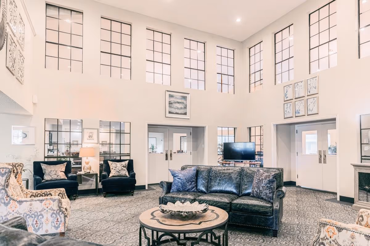 A spacious and bright senior living facility common area with high ceilings and large windows. The room features a black leather sofa, patterned armchairs, a round wooden coffee table, and two dark blue armchairs with decorative pillows. There is a flat-screen TV on a stand, framed artwork on the walls, and a patterned carpet covering the floor.